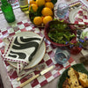 A meal table with a checkered cloth, lemons, wine, water bottles, salad, bread, and The Poppy Tile Venice Napkin by Good Juju Homeware enhances the vibrant Poppy Tile print tableware.