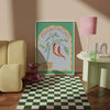 The Sunday Chilli Wall Art by Good Juju Homeware, depicting vibrant chili peppers, leans against a wall beside a beige sofa. Nearby are a pink lamp, green table with a glass, and checkered rug on parquet flooring, evoking Mediterranean flair.