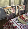 Outdoor dining setup with two chairs featuring patterned cushions, and a table covered with a matching checkered tablecloth set with plates. Sunlight casts shadows across the scene.