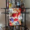 A colorful tea towel with abstract design hangs on a bar cart filled with various bottles and a crystal decanter.