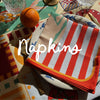 Striped red and white napkin on a plate, surrounded by an orange, cutlery, and glassware. Text overlay reads "Napkins.