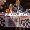 Colorful tablecloth displayed on a table with fruits, a pitcher, and decorative items, with the word "Tablecloths" written across the image.