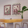 A modern room features a wooden table, two chairs, and framed "Tropical Flowers in a Glass Vase Venice Wall Art" by Good Juju Homeware. The artwork’s floral design enhances positivity, while a green plant adds elegance to the table.