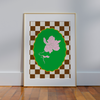 The Framed Magnolia Florence Wall Art by Good Juju Homeware, with a pink flower on a green backdrop and a brown and white checkered border, elegantly leans against a white wall on a wooden floor, exuding nature-inspired sophistication.