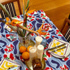 The table is elegantly adorned with a Poppies All Around Milan Tablecloth from Good Juju Homeware, perfect for a 6-8 seater setup. It features geometric napkins, tropical flowers in a vase, candles, fresh oranges, and blue glasses—all creating an inviting atmosphere for any gathering.