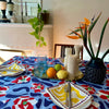 A vibrant Poppies All Around Milan Tablecloth by Good Juju Homeware graces the 6-8 seater dining table. The patterned plates, utensils, and glasses add charm. A fruit tray takes center stage, surrounded by decorative vases with candles and a plant.