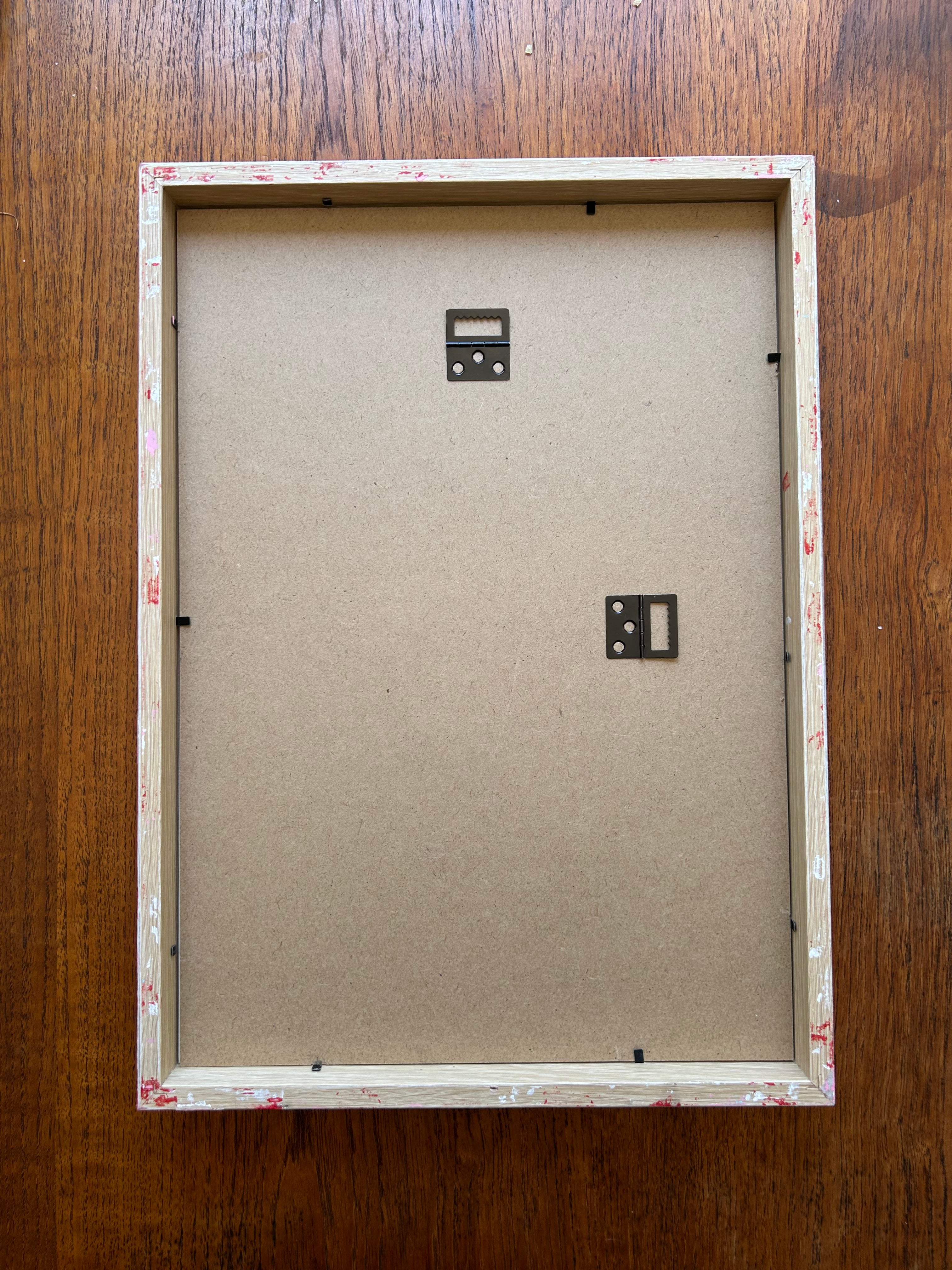 The A3 Dark Turquoise and Light Green Painted Frame by Good Juju Homeware rests on a wooden surface, featuring hand-painted stripes, metal hanging brackets, a cardboard backing, and small paint stains on its edges.