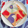 The Poppies All Around Rome Napkin by Good Juju Homeware graces a white plate, enhancing the tableware collection with its vibrant abstract patterns. Complemented by similar designs on a tablecloth, it brings creativity and style to your setting.