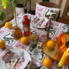 A table with The Poppy Stripe Venice Napkin from Good Juju Homeware, set with exquisite tableware and Aperol, is surrounded by vibrant plants. Oranges, candles, and glasses complete the inviting scene.