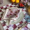 A table setting with The Poppy Tile Venice Napkin by Good Juju Homeware, a fruit centerpiece, elegant tableware, bowls of bright lemons and tomatoes, glasses, a bottle, and a candle on a checkered tablecloth.