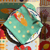 A plate with The Sunday Pesce Napkin by Good Juju Homeware showcases an orange fish design. Behind it, a tablecloth displays a colorful Mediterranean pattern with food illustrations. A fork and knife sit to the right, hinting at an Italian feast.