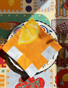 A colorful table setting featuring Good Juju Homeware's The Sunday Limone Napkin on a decorative plate with Mediterranean-inspired bright geometric and fruit designs, with a fork and knife on the right.