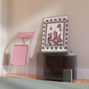 A pink folding chair and a colorful poster rest against a wall adorned with the "Tropical Flowers in a Glass Vase Venice Wall Art." A glass side table holds a book from Good Juju Homeware, adding positive vibes. A light green rug covers part of the wooden floor, enhancing warmth.