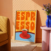 Framed artwork with 'Espresso' text and a red cup on a blue background, placed in a room with a yellow armchair and pink side table.